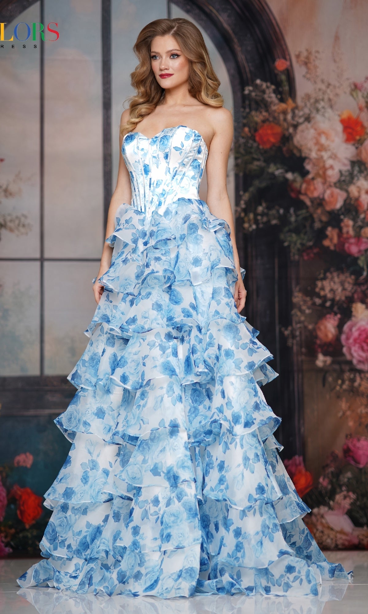 A woman stands before a floral arched window, wearing the Colors Dress 3836—a strapless, floor-length ruffle prom gown by Colors Dress. The blue and white floral pattern and her long wavy hair create an elegant, confident look.